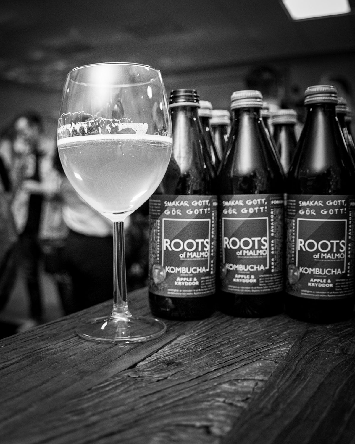 A glass next to bottles of Roots of Malmö kombucha on a wooden bar