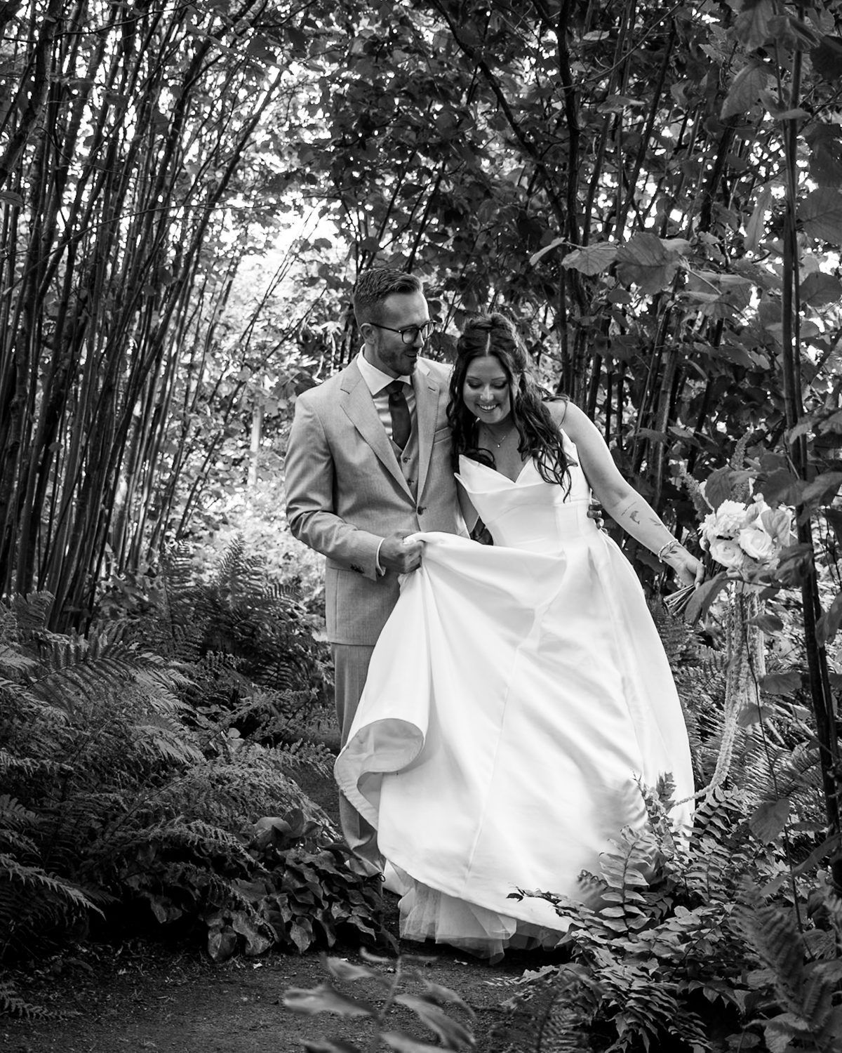 Bride and groom laughing on a fern-lined garden path, the groom holding up her dress